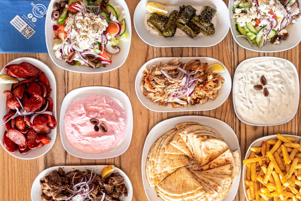 Flat lay photo of assortment of Greek food dishes including chips, pita bread, meat packs, lab, chicken, sauce, salads and chorizo. All plates are sitting on timber table.