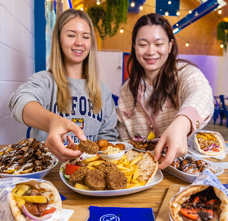 Two happy girls in a Souvlaki GR restaurant sitting side by side reaching for a share plate of Greek Food, with an assortment of Greek food dishes surrounding them.
