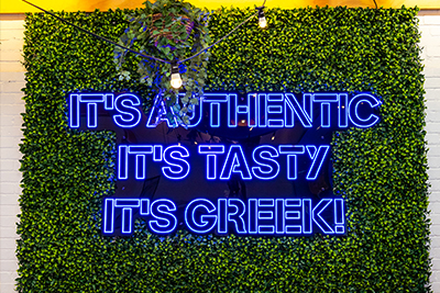 Photo of a tagline neon sign with the words "It's Authentic, it's tasty, it's Greek!" on a decorative grass wall