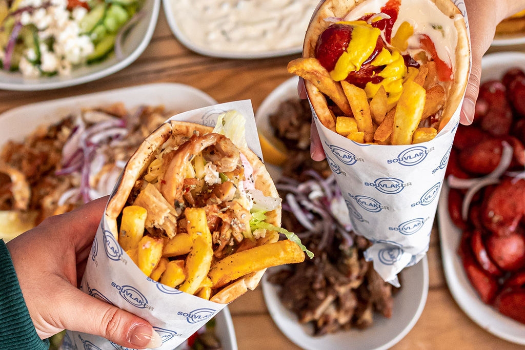 two hands holding a souvlaki each, with Greek food in the background