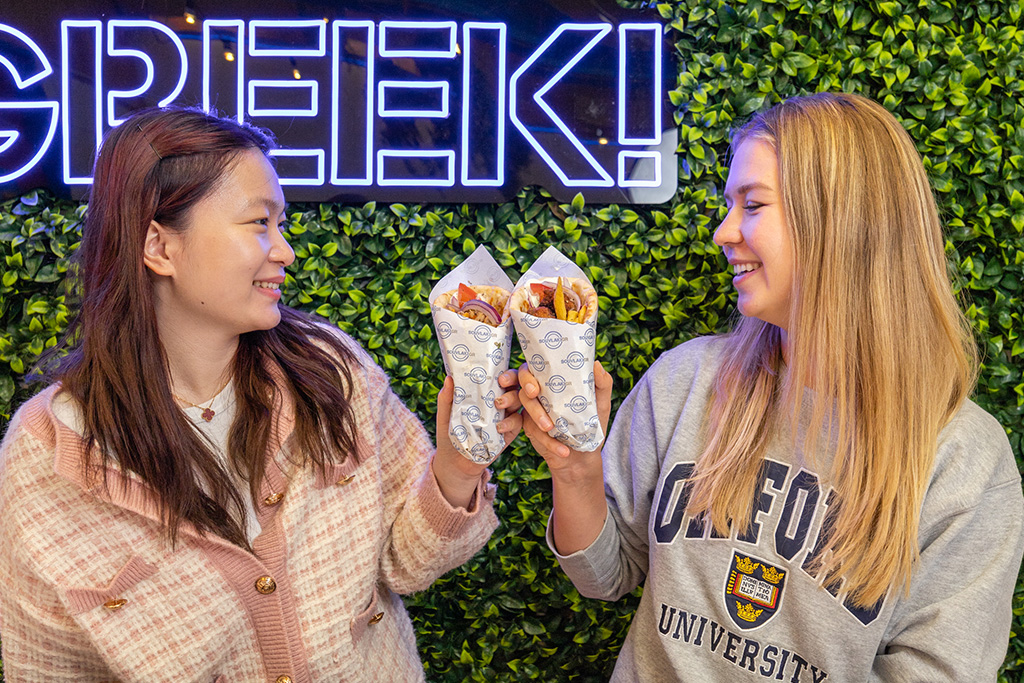 Two girls smiling holding a souvlaki in their hands and making them touch.