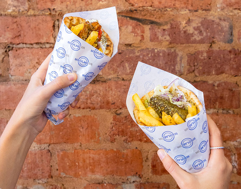 Two hands holding up a souvlaki in each with an exposed brick wall in the background