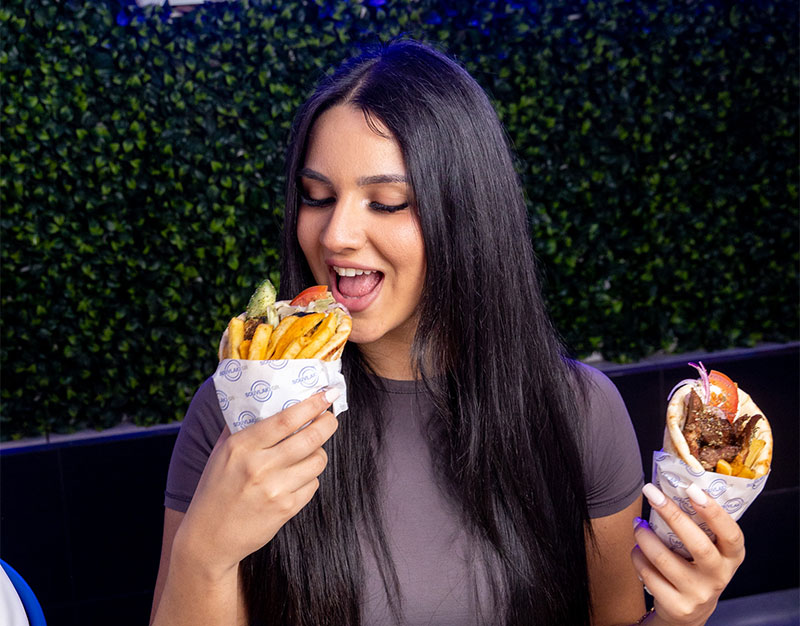 Happy lady holding a Souvlaki in each hand, holding on up to her mouth ready to take a bite