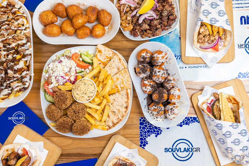 Flay lay photograph of an assortment of Souvlaki GR Greek food, including souvlakia, meat, share plates, HSP's and desserts all sitting on a table.