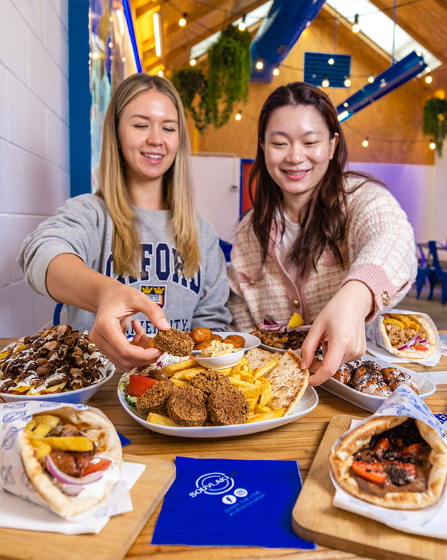 Two happy females sitting side by side enjoying a share plate inside of a Souvlaki GR restaurant. They are surrounded with other Greek food items such as souvlakia, HSPs and desserts.