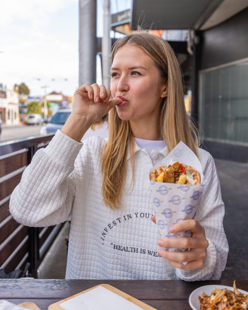 Girl holding a souvlaki sitting out the front of a Souvlaki GR restaurant licking sauce of finger